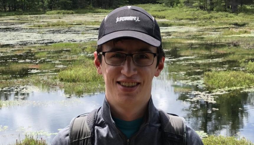 A man with a black shirt, with glasses and hat. Behind him, is greenery. 