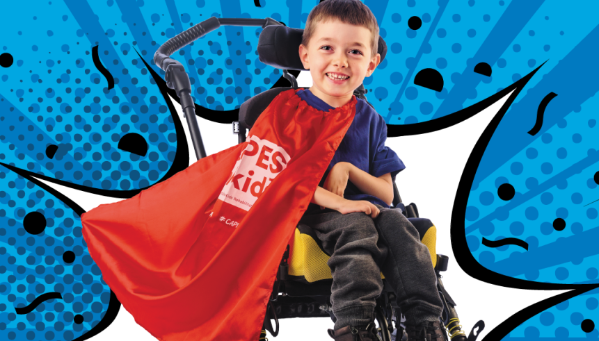 Young boy sitting in a wheelchair wearing a red cape. Photo on a blue background.