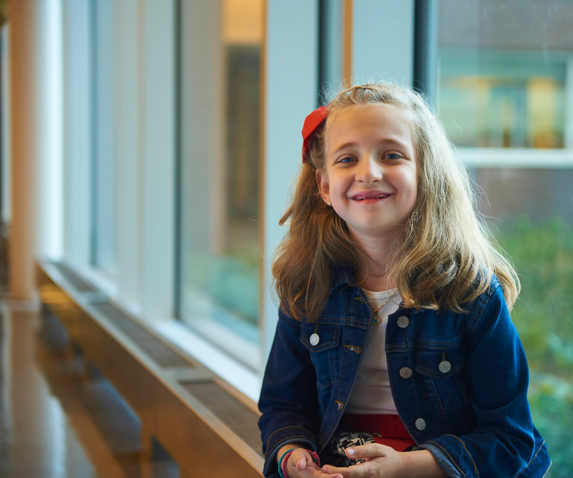 little girl with light skin tone and long blonde hair with a red bow in it wearing a jean jacket