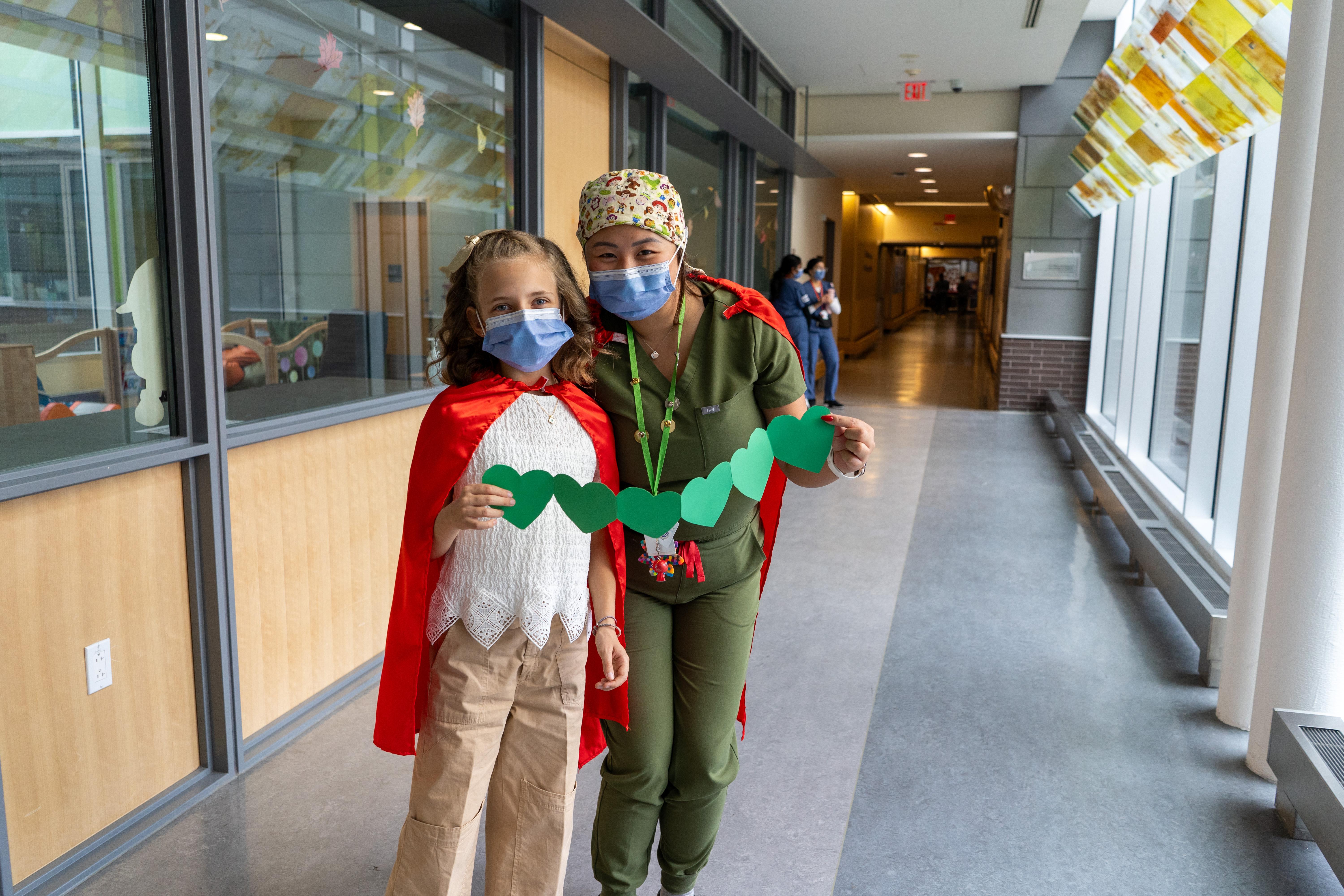 A young girl with blonde hair and a nurse standing side by side wearing red capes and holding green cutout paper hearts