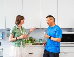 Two young adults having a conversation in a kitchen, standing in front of some cabinets 