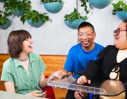 Three young adults, one of whom uses a wheelchair, sitting at a table, talking and laughing 