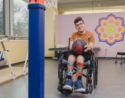 A teenager who uses wheelchair holding a basketball in their lap and smiling. In front of them is a basketball hoop 