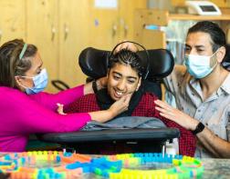 Two adults are helping a teenager in a wheelchair try on a BCI headset
