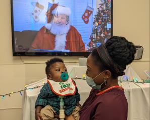 Parent, Julia, holding her child (and Holland Bloorview client), Thomas in front of a TV screen featuring Santa.