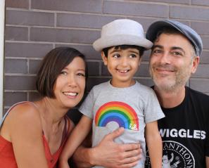 A woman with short brown hair is smiling and hugging her young son. The young boy is smiling and wearing a grey t-shirt with a rainbow on it. His father is kneeling beside him and holding him. He is wearing a black t-shirt and a baseball cap.