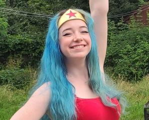 An adult with light skin tone and long blue hair wearing a gold crown-shaped hairband and a red tank top.