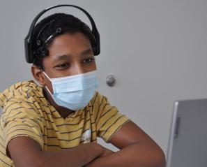 A boy with brown skin tone and black hair in a striped shirt, wearing a mask and headphones seated in front of a computer