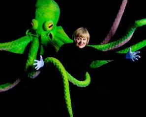 Woman in black smiling surrounded by giant bright green octopus and its tentacles.