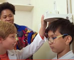 A child pretending as doctor to check the height of another child, an adult is standing behind them.