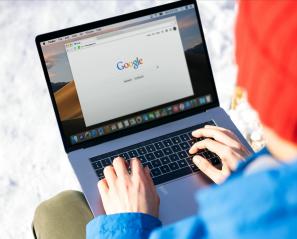 Person wearing a blue hoodie and orange toque is sitting with a grey laptop open and browsing on the Internet.