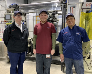 Alex stands in between two other adults in front of some factory equipment
