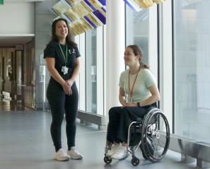 Two women smile beside floor to ceiling window, one in a wheelchair