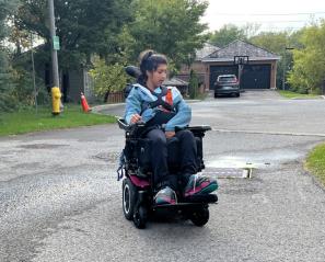 Teen with long dark hair drives power wheelchair in suburban neighbourhood.