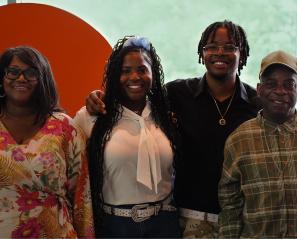 Photograph of four people standing side by side in front of a large orange circular backdrop with a green blurred background. They are wearing casual clothing, 