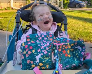 Young girl with oxygen prongs and huge smile sits in wheelchair with backpack on tray.