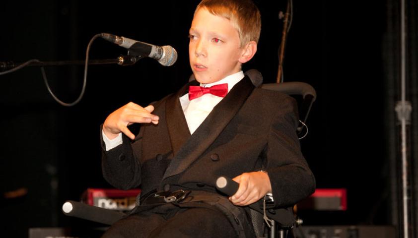 A young person wearing a suit and red bow tie. They are speaking into a microphone.