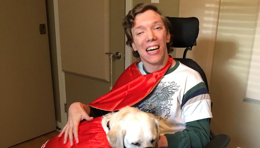 A young adult wearing a red cape, smiling and using a wheelchair. They are cradling a large white dog.