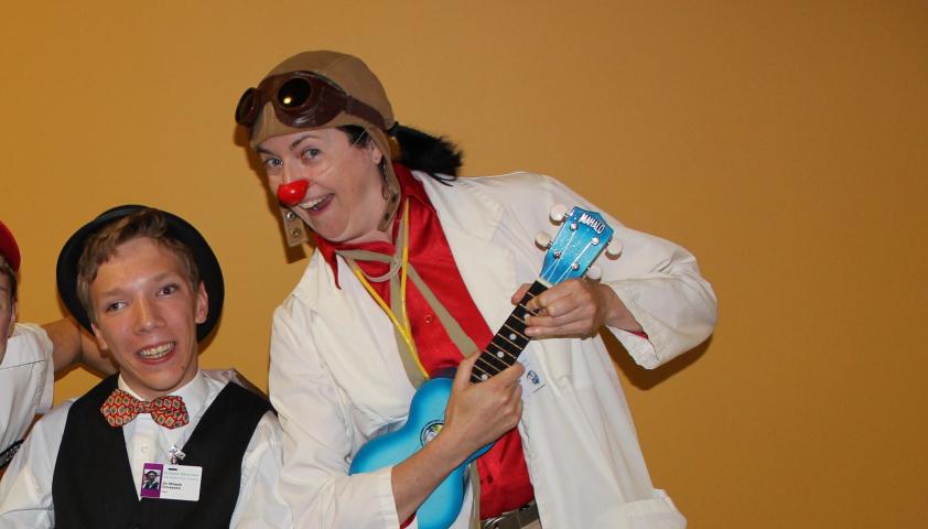 A young person wearing a top hat, tie and a vest. They are smiling and posing next to an adult wearing a clown nose and playing a blue ukulele.