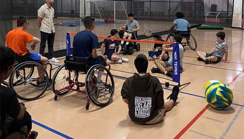 A group of teeangers with wheelchair listening to a basketball coach in a basketball court