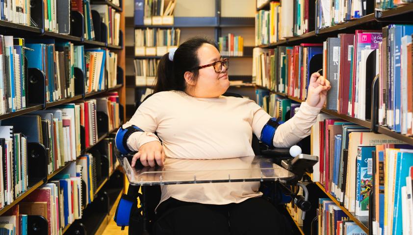 A young woman who uses a powerchair reaches for a book on a library shelf