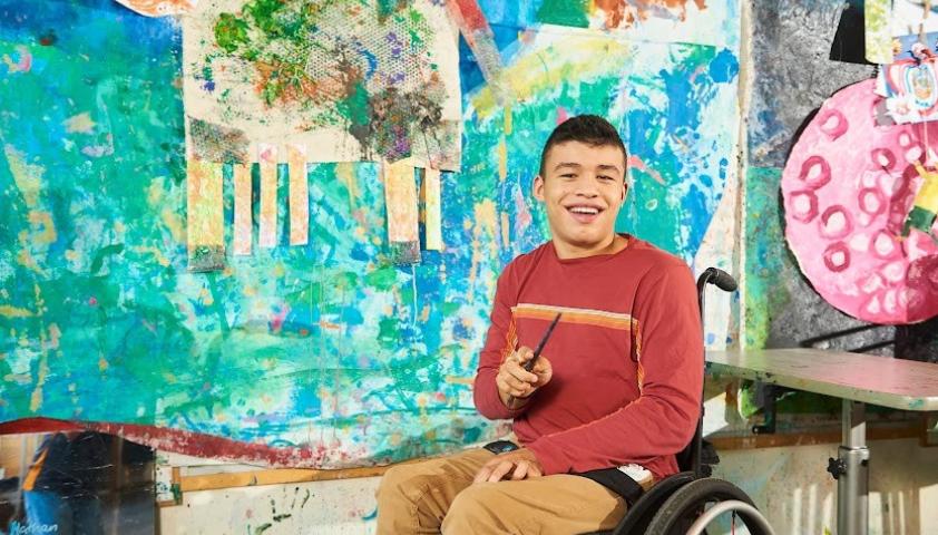 A young person who uses a wheelchair smiles and holds a paintbrush in front of a colourful mural.