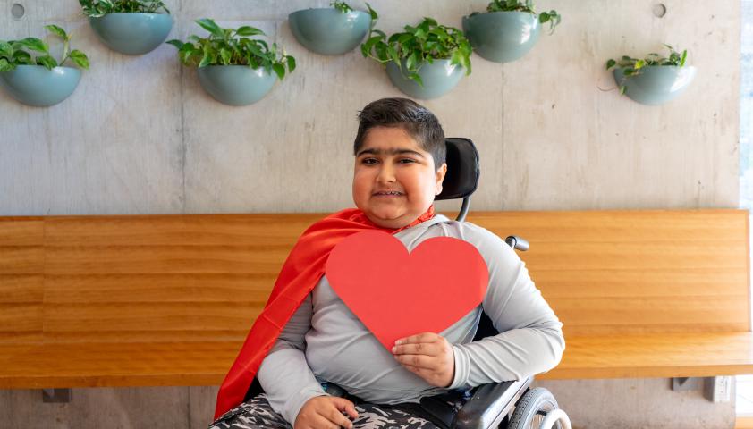 A young boy wearing a red cape and holding a big red paper heart.