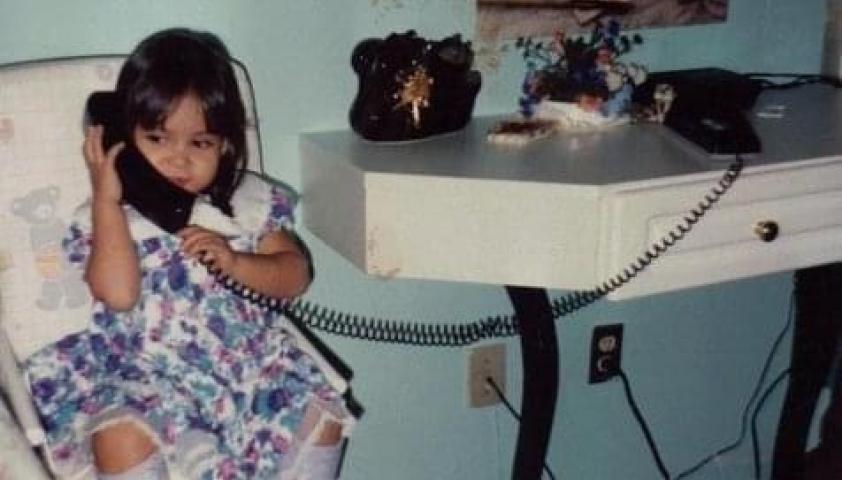 Dennisse as a child, sitting in a high chair and holding a landline phone to her ear