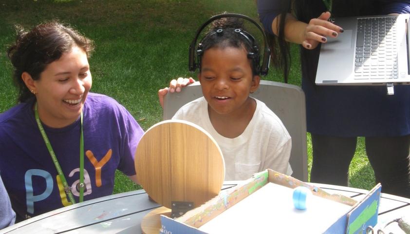 A child wearing a BCI headset to paint outside. An adult is kneeling by the child, and another adult is standing with a laptop.