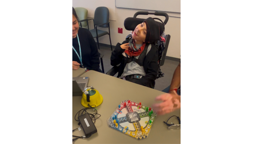 A child in a wheelchair, sitting at a table. There is a board game and a dice roller on the table.