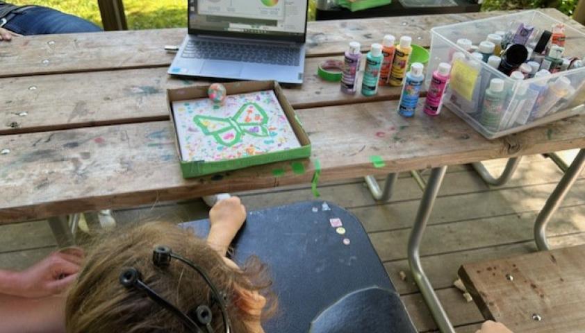 An outdoor table with a laptop, paints, and a painting project. A child is wearing a BCI headset, and is using the BCI to move a small ball with paint on it, to create a painting of a butterfly.