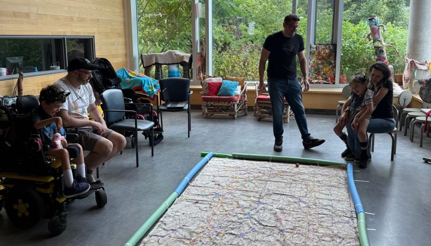 Two children are painting together. The children are wearing BCI headsets to control robotic balls dipped in acrylic paint. The painting is large and covers the floor. One child is in a wheelchair with his dad sitting next to him. The second child is sitting on her mom’s lap, with her dad standing beside them.