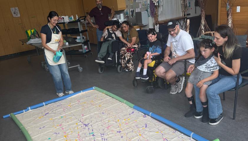 Three children are painting together. The children are wearing BCI headsets to control robotic balls dipped in acrylic paint. The painting is large and covers the floor. Two children are in wheelchairs and are sitting beside their parents, and one child sits on their mom’s lap. A clinician is standing and pouring paint onto the painting.