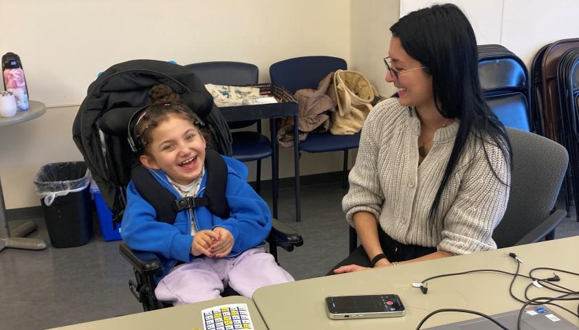 A child and adult sitting next to each other, playing a game. The child is in a wheelchair and wearing a BCI headset.