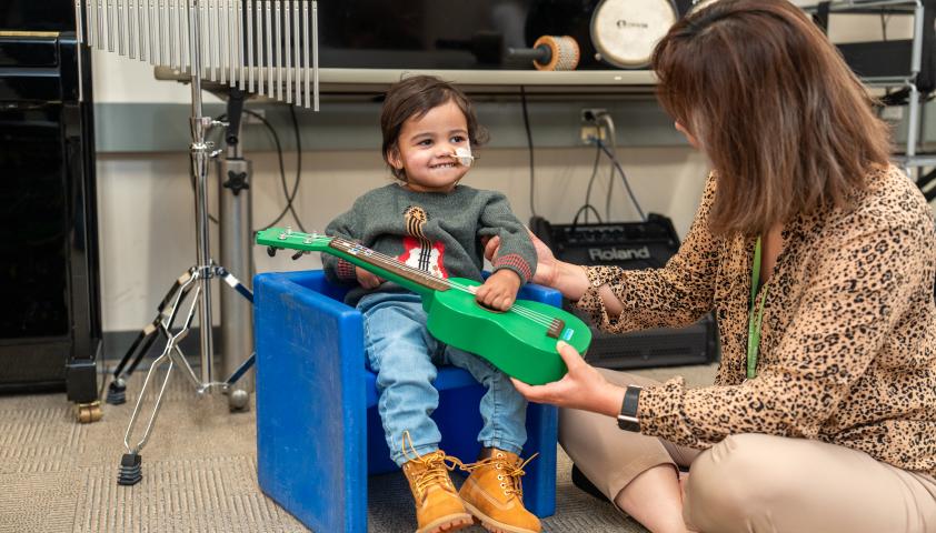 A young boy and a music therapist strum a green guitar