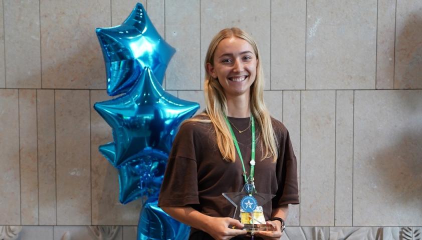 Woman with blond hair and wearing a brown blouse holds an award and smiling.