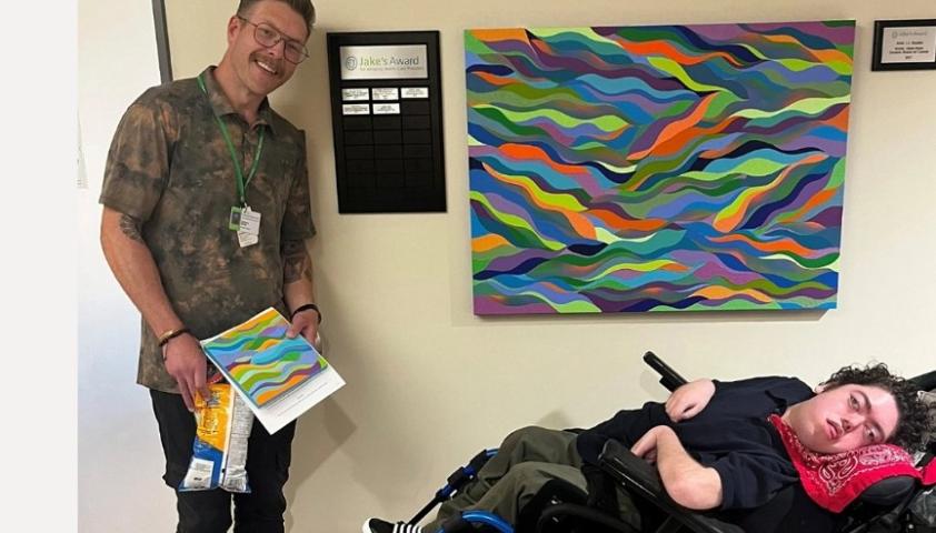 Adult male stands with painting in his hand next to a similar painting on the wall and a youth in a wheelchair.