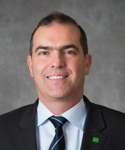 Man with dark hair wearing a black blazer, white shirt, and a blue-striped tie.
