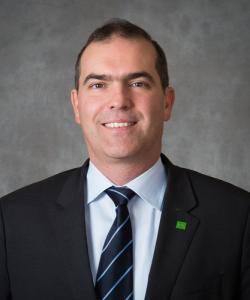 Man with dark hair wearing a black blazer, white shirt, and a blue-striped tie.