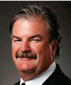 Man with brown hair and a moustache wearing a black blazer and striped tie