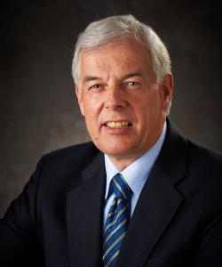 Man with gray hair wearing a dark blazer, light blue shirt, and a blue-striped tie