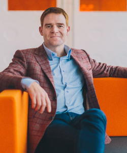 Man with short brown hair wearing a light blue shirt and plaid blazer sitting on a bright orange couch.