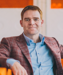 Man with short brown hair wearing a light blue shirt and plaid blazer sitting on a bright orange couch.