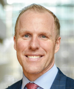 A smiling man with short blond hair and blue eyes. Wearing a dark blue plaid blazer, a light blue dress shirt, and a red patterned tie