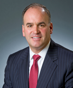 A smiling man with sort dark hair wearing a dark blue pinstriped blazer, white dress shirt, and red patterned tie