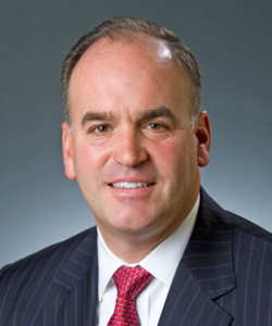 A smiling man with sort dark hair wearing a dark blue pinstriped blazer, white dress shirt, and red patterned tie