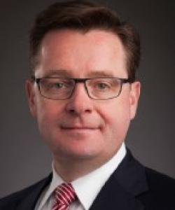 Man with short dark hair and black rectangular glasses. He's wearing a black blazer, white dress shirt, and a red and white striped tie