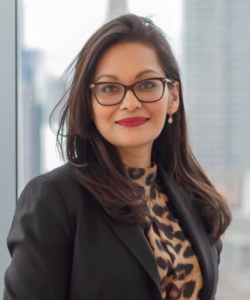 Woman with long dark hair and red lipstick. She is wearing glasses, a leopard print shirt, and a black blazer.