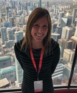 A woman with shoulder length hair wearing a black shirt, she is wearing a red lanyard with a name tag. In the background, outside the window, there are buildings. 