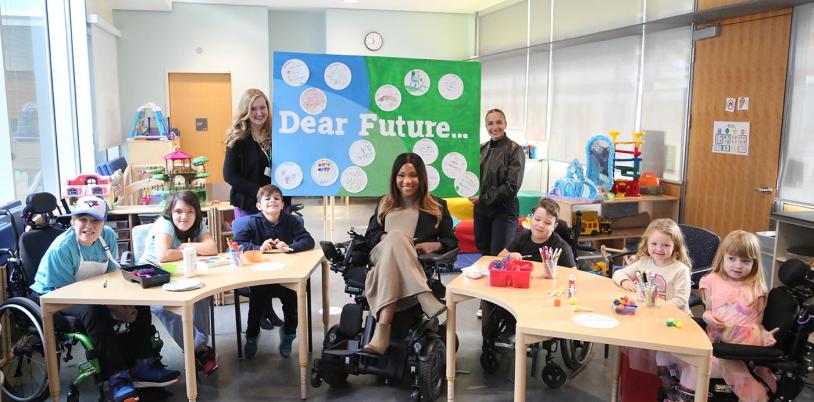 Children, teenagers and adults sitting and standing at a table of craft supplies. Two adults hold a green and blue banner that reads, 'Dear Future.'